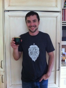Founder of the Forest City Beer Fest, Aaron Brown, holding a glass of beer with "Forest City Beer Fest" on the a green outline of a leaf. His navy t-shirt also has the logo, but the leaf is filled in white. 