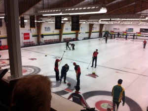 The Fanshawe Men's Curling team on the ice, with NAIT. Person is watching from the balcony
